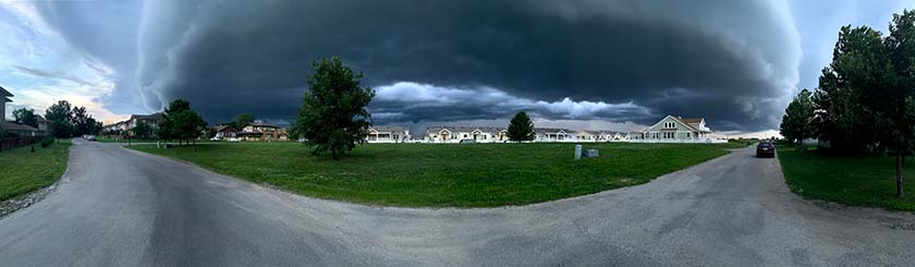 Panaramic photo of a neighborhood street with a large dark cloud in the sky.